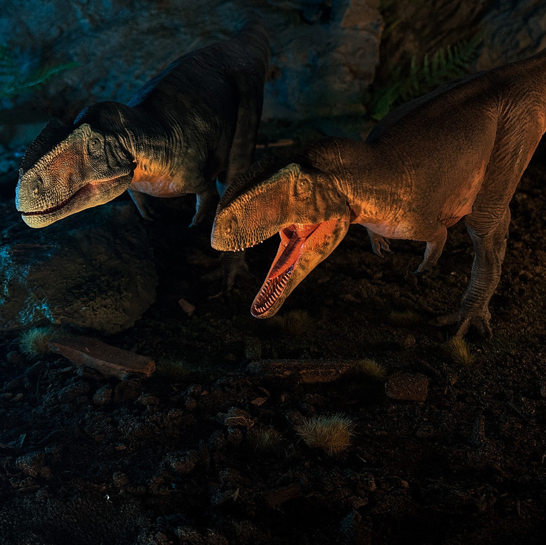 Two GIGANOTOSAURUS dinosaur models in a dark, rocky setting with glowing blue light.